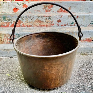 May include: A large, copper pot with a single iron handle. The pot is aged and has a rustic appearance. It is sitting on a concrete surface.
