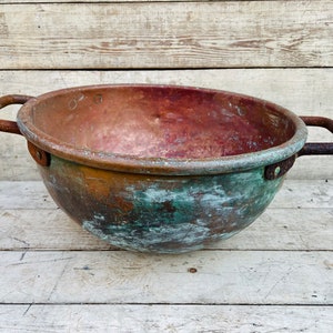 Może przedstawiać: Duża, antyczna miedziana miska z dwoma uchwytami. Miska ma zieloną patynę i jest lekko wgnieciona.