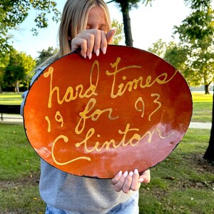 May include: Oval-shaped, orange-colored decorative plate with the words "hard times for Clinton 1993" in gold script. The plate has a dark border and is held in front of a person in a park setting.