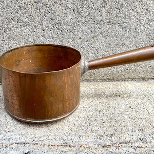 Może przedstawiać: Stary miedziany garnek z długim uchwytem. Garnek ma zużytą, starą patynę i idealnie nadaje się do rustykalnego wystroju kuchni.