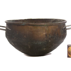 Może przedstawiać: Duży, antyczny miedziany garnek z dwoma metalowymi uchwytami. Garnek jest zużyty i ma patynę. Mała puszka piwa Krueger's Finest Beer znajduje się na pierwszym planie w celu porównania rozmiarów.