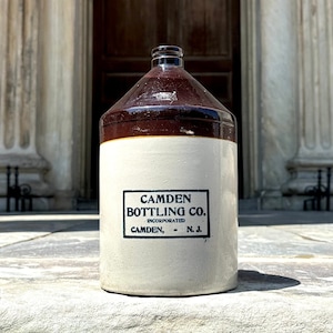 May include: A vintage brown and white ceramic jug with a brown lid. The jug has a label that reads "CAMDEN BOTTLING CO. INCORPORATED CAMDEN, - N.J."