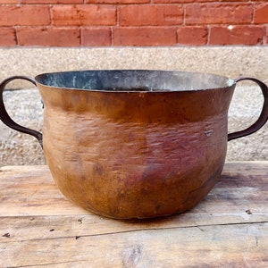 Könnte beinhalten: Ein großer Kupfertopf mit zwei Griffen. Der Topf hat eine gehämmerte Oberfläche und ist leicht verbeult.