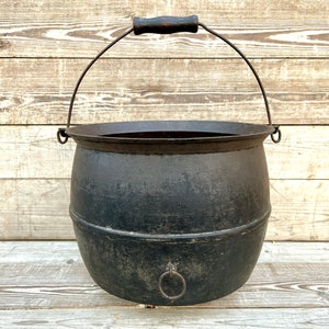 May include: A black cast iron cauldron with a wooden handle and a metal ring on the side. The cauldron is sitting on a wooden surface.