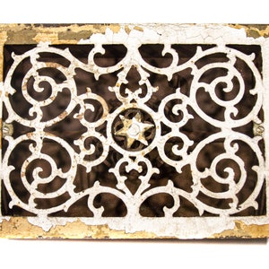 Puede incluir: Una rejilla de ventilación de hierro fundido vintage con un diseño decorativo y ornamentado. La rejilla está pintada de blanco, con algunas áreas de la pintura descascarada, revelando el óxido debajo.