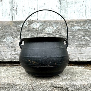 Puede incluir: Un caldero de hierro fundido negro con un asa de alambre. El caldero está sentado sobre una superficie de madera.
