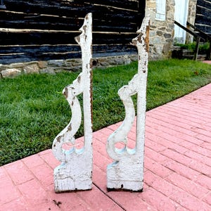 May include: Two antique, white-painted wooden architectural brackets with a distressed finish. The brackets have a scroll design and are weathered, showing signs of age and use. They are placed on a pink brick surface with a green lawn in the background.