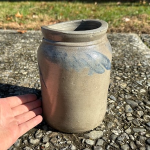 Puede incluir: Un tarro de gres cilíndrico con una abertura ancha y un borde ligeramente curvado. El exterior es de color marrón moteado con pinceladas azules. El tarro parece hecho a mano.