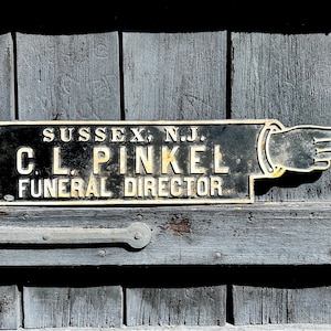 Puede incluir: Un letrero de metal negro y blanco con una mano señalando y el texto "SUSSEX, N.J. C.L. PINKEL FUNERAL DIRECTOR".