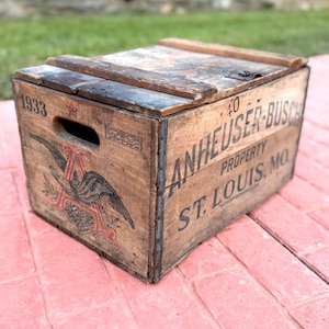 May include: A vintage wooden Anheuser-Busch crate with a hinged lid. The crate displays the Anheuser-Busch logo with an eagle and the text "ANHEUSER-BUSCH PROPERTY ST. LOUIS, MO." The year "1933" is printed on the side.