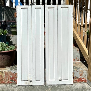 May include: A pair of white wooden shutters with a simple design. The shutters are closed and have a knob on each side.