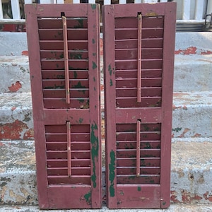 Puede incluir: Dos contraventanas de madera roja desgastada con lamas horizontales y soportes verticales. Las contraventanas muestran signos de envejecimiento, con pintura descascarillada y detalles verdes, sugiriendo una estética vintage o rústica. Para la decoración del hogar.