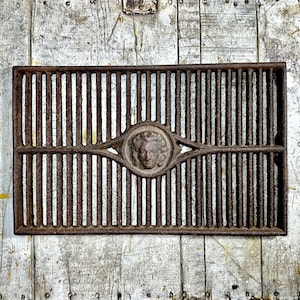 May include: A rusty cast iron grate with a decorative design featuring a face in a circle. The grate is rectangular and has vertical bars.