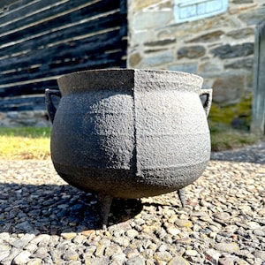 May include: A black cast iron cauldron with three legs and two handles. The cauldron is sitting on a bed of small stones.
