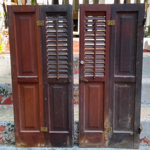May include: Two antique wooden shutters with louvered panels and decorative trim. The shutters are painted in a dark brown color and have brass hinges.