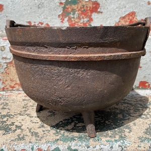 May include: A rustic, cast iron pot with three legs and two handles. The pot is dark brown with a worn, aged appearance.
