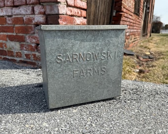 Op de afbeelding: Een vintage gegalvaniseerde metalen container met de woorden "SARNOWSKI FARMS" erop gegraveerd. De container staat op een grijs, getextureerd oppervlak.