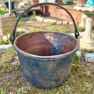 May include: A large, antique copper cauldron with a dark, weathered exterior and a prominent iron handle. The interior shows a mix of copper and verdigris, suggesting age and use. The cauldron sits on grass, hinting at outdoor use.