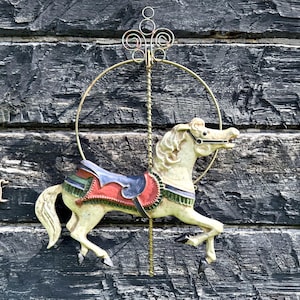 May include: A vintage carousel horse figurine with a cream-colored body, a red and blue saddle, and a gold-colored pole and decorative top. The horse is set against a weathered, dark wood background.
