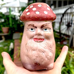 May include: A quirky, hand-painted mushroom ornament with a red cap and white spots. The mushroom features a face with blue eyes, a red mouth, and a long, white beard. The ornament is held in a person's hand.