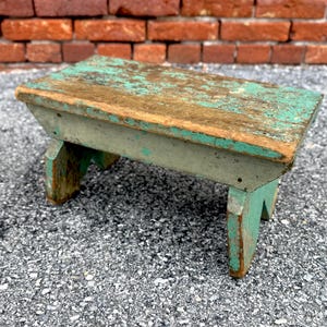 Könnte beinhalten: Ein kleiner, rustikaler Holzhocker, der in einem verblassten Teal-Grün gestrichen ist. Der Hocker hat eine einfache, rechteckige Oberseite und zwei abgewinkelte Beine. Das Holz weist Gebrauchsspuren auf, die ihm einen Vintage-Look verleihen.