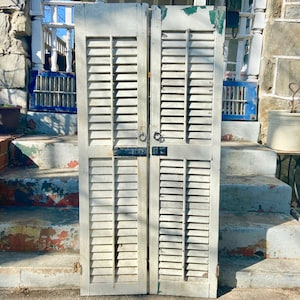 Puede incluir: Un par de contraventanas de madera blanca con herrajes de metal negro. Las contraventanas están cerradas y tienen un aspecto desgastado. Están colocadas en un conjunto de escalones de hormigón.