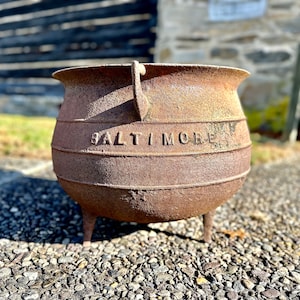 May include: A weathered, rusty cast iron pot with three legs and a handle. The word "BALTIMORE" is embossed on the side. The pot is outdoors on a gravel surface, with a blurred background of a wooden structure.