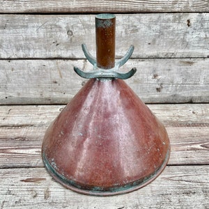 Puede incluir: Un embudo de color cobre con forma cónica y parte superior cilíndrica. El embudo tiene una base ensanchada y tres asas curvas cerca de la parte superior. El metal muestra signos de edad y desgaste, con pátina y algo de oxidación verde.