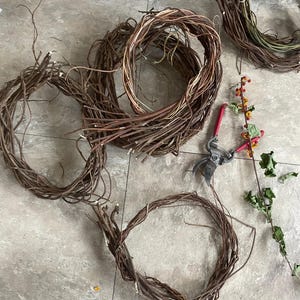 May include: Several unfinished grapevine wreaths in various stages of completion are arranged on a light-colored surface. A pair of red-handled pruning shears and a sprig of berries and leaves are also present, suggesting a crafting project.