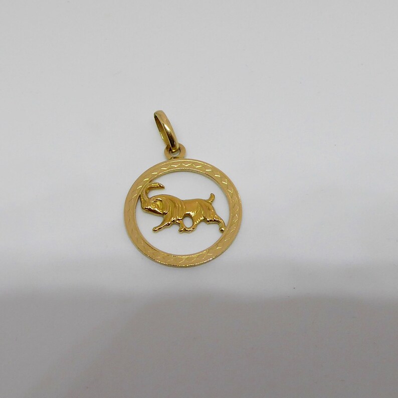 May include: Gold pendant featuring a circular design with a detailed outline and a depiction of a goat inside.