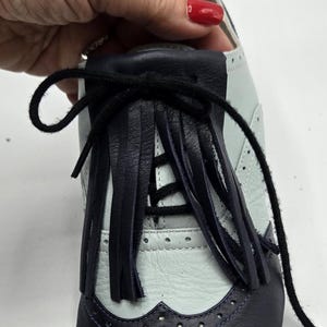 May include: A two-tone leather shoe with a navy blue toe and heel, and a light blue upper. The shoe features a decorative fringe and black laces. The design includes brogue detailing and a classic Oxford style.