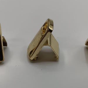 May include: Three gold-colored metal clasps. The clasps have a rectangular shape with a loop at the top. One clasp has a black square base. The clasps are likely used for bags or other accessories.