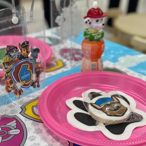 May include: A Paw Patrol themed party setup with a pink plate holding a cookie featuring Chase. The table setting includes a Paw Patrol emblem, a juice bottle with a Marshall figurine, and clear plastic bags.
