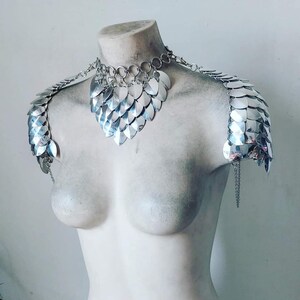 May include: Silver chainmail shoulder armor and necklace set. The set is made of interlocking metal scales and features a heart-shaped neckline.