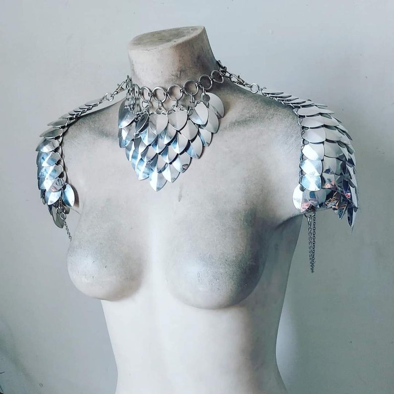 May include: Silver chainmail shoulder armor and necklace set. The set is made of interlocking metal scales and features a heart-shaped neckline.