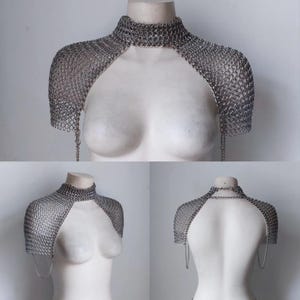 Puede incluir: Una hombrera de cota de malla plateada con cuello alto. La cota de malla está hecha de pequeños anillos metálicos entrelazados. La pieza cubre los hombros y la parte superior del pecho, con cadenas colgantes.