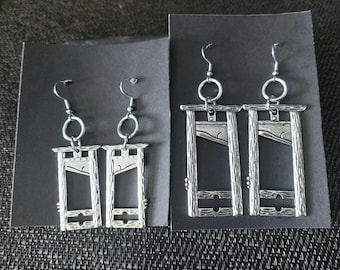 Puede incluir: Dos pares de pendientes de guillotina plateados. Cada pendiente presenta un diseño de guillotina detallado con una cuchilla y un marco texturizado de madera.