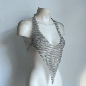 May include: A silver chainmail halter top with a triangular design. The top is displayed on a white bust, showcasing the intricate metal links and the garment's shape. The halter top is a unique piece of body jewelry.