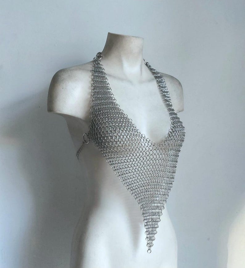 May include: A silver chainmail halter top with a triangular design. The top is displayed on a white bust, showcasing the intricate metal links and the garment's shape. The halter top is a unique piece of body jewelry.