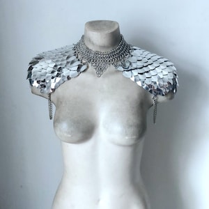 Puede incluir: Una hombrera plateada con un cuello de cota de malla y hombreras en forma de escamas. La pieza se exhibe sobre un busto blanco, con detalles de cadena colgante. El diseño evoca una estética medieval o fantástica.