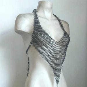 May include: A silver chainmail halter top with a triangular design. The top is displayed on a white mannequin, showcasing the intricate metalwork and open weave. The halter straps and back closure are also made of chainmail.