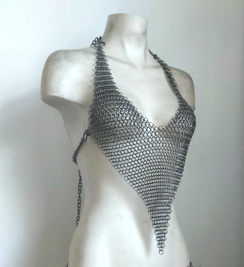 May include: A silver chainmail halter top with a triangular design. The top is displayed on a white mannequin, showcasing the intricate metalwork and open weave. The halter straps and back closure are also made of chainmail.