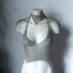May include: A silver-toned chainmail halter top featuring a triangular neck piece connected by thin metal chains to a circular ring at each shoulder. The garment is constructed from small interlocking metal rings forming a mesh pattern.