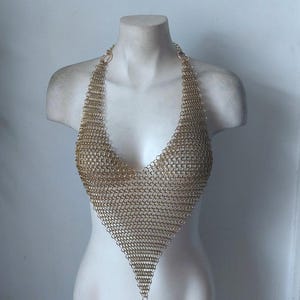 May include: A gold-colored chainmail halter top displayed on a white mannequin bust. The top is constructed of small, interlocking metal rings, forming a V-neck and a pointed bottom. The halter straps connect to the top with circular rings.