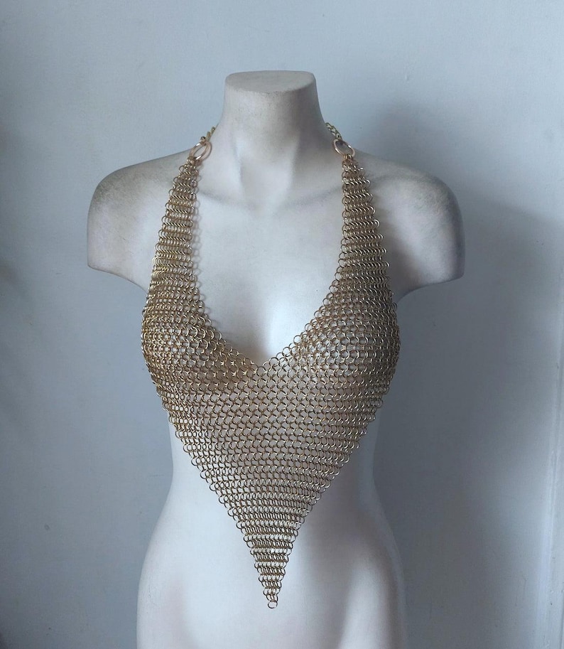 May include: A gold-colored chainmail halter top displayed on a white mannequin bust. The top is constructed of small, interlocking metal rings, forming a V-neck and a pointed bottom. The halter straps connect to the top with circular rings.