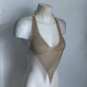 May include: A gold-colored chainmail halter top with a plunging V-neck design. The top is made of small, interlocking metal rings, creating a flexible, textured fabric. The halter straps are attached to the top with small rings.