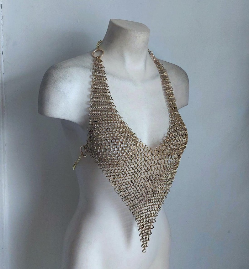 May include: A gold-colored chainmail halter top with a plunging V-neck design. The top is made of small, interlocking metal rings, creating a flexible, textured fabric. The halter straps are attached to the top with small rings.