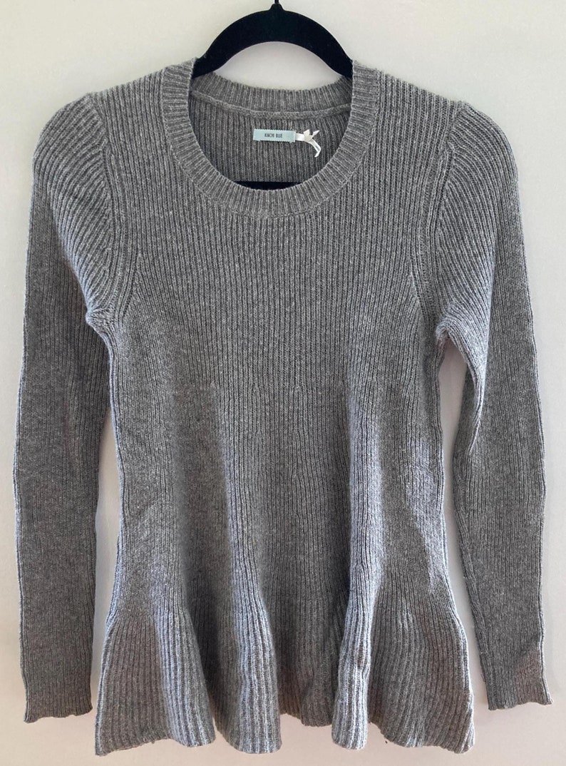 May include: Gray ribbed sweater with a ruffled hem and long sleeves. The sweater has a round neckline and a small white tag with the text "A New Day" on it.