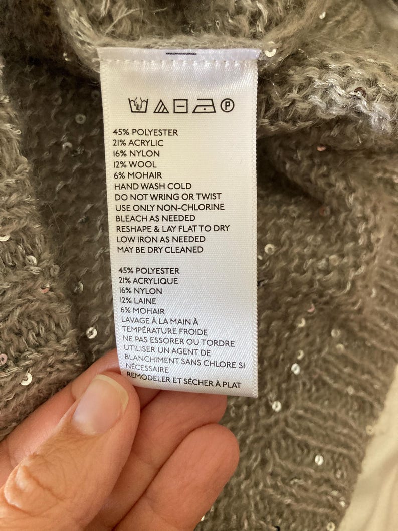 Op de afbeelding: Een wit waslabel met zwarte tekst met details over de stofsamenstelling en wasinstructies voor een kledingstuk. Het label vermeldt: 45% Polyester, 21% Acryl, 16% Nylon, 12% Wol, 6% Mohair. Handwas koud, niet wringen of draaien, alleen chloorvrij bleekmiddel gebruiken indien nodig, in vorm brengen en plat laten drogen, strijken op lage temperatuur indien nodig, chemisch reinigen mogelijk. 45% Polyester, 21% Acryl, 16% Nylon, 12% Wol, 6% Mohair. Handwas koud, niet wringen of draaien, alleen chloorvrij bleekmiddel gebruiken indien nodig, in vorm brengen en plat laten drogen, strijken op lage temperatuur indien nodig, chemisch reinigen mogelijk.