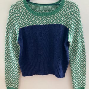 May include: A green and white patterned sweater with a blue cable knit panel in the center. The sweater has long sleeves and a round neckline.
